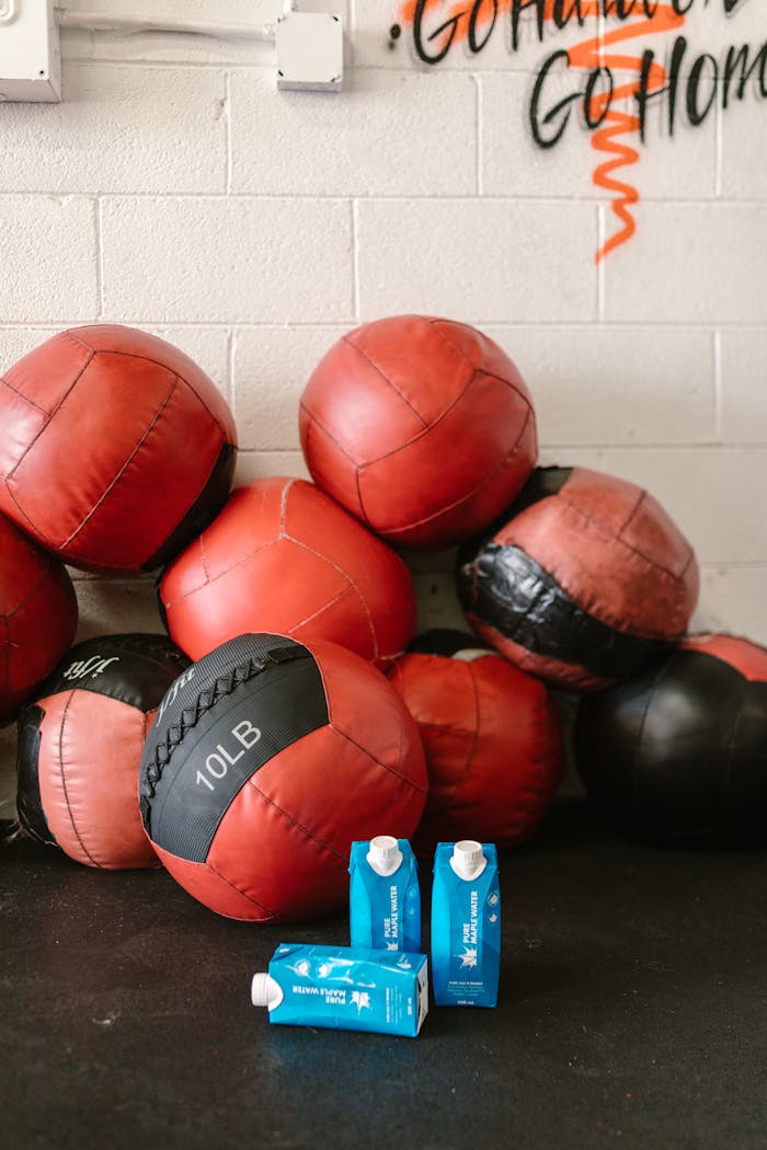 Red and black medicine balls stacked in a gym setting with two blue hydration cartons on a dark floor.